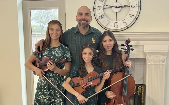Dr. Michael Cerean with children holding string instruments, grouped near a fireplace and decorative wall clock.